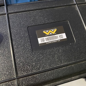 Op de afbeelding: Zwarte kunststof koffer met een tamper-evident tag. De tag heeft een Weyland-Yutani Corp logo met de tekst "Building Better Worlds".