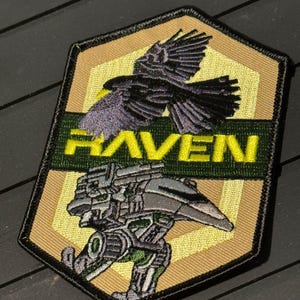 May include: Embroidered patch with a raven in flight above the word "RAVEN" in yellow. Below the text is a detailed illustration of a mechanical figure. The patch has a tan, green, and black color scheme.