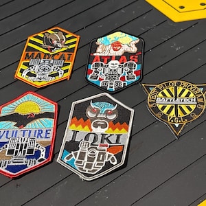 May include: Five embroidered patches with various designs and text. The patches feature images of robots, a sun, and text such as "MADCAT", "ATLAS", "VULTURE", "LOKI", and "BATTLETECH". The patches are in various colors, including yellow, blue, red, and black.