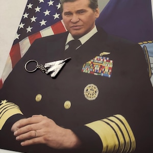 May include: A portrait of a man in a navy uniform with gold stripes and insignia. An American flag is in the background. A silver airplane keyring is in the foreground.