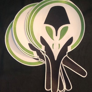 Puede incluir: Cuatro parches bordados blancos, verdes y negros con un diseño de cara alienígena.