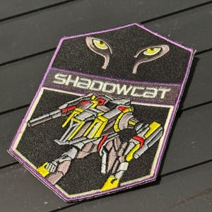 May include: Embroidered patch featuring a robotic figure with yellow, red, and gray accents, labeled "SHADOWCAT." The patch has a black background with a purple border and the image of cat eyes at the top.