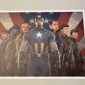 Peut inclure: Un groupe de sept hommes en uniforme militaire se tiennent devant un drapeau américain. L'homme au centre porte un costume de Captain America bleu et rouge. Les autres hommes portent des uniformes de différentes nuances de brun et de vert.