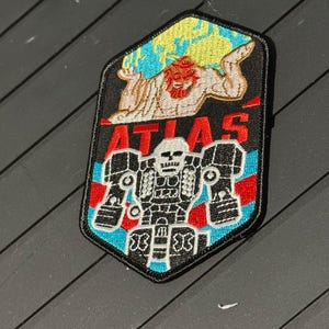May include: Embroidered patch with a black border, featuring a red-haired Atlas figure holding a globe, with the word "ATLAS" in red. Below, a white and black robot design is set against a blue and red striped background.
