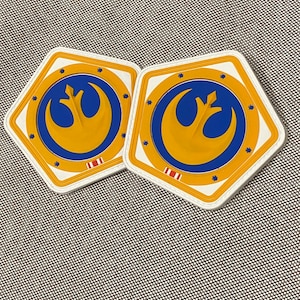 May include: Two white, hexagonal-shaped patches with a blue and gold Rebel Alliance symbol. The symbol features a white bird with wings outstretched, a blue circle around it, and a gold ring around the circle. The patches have a white border and a red and white stripe at the bottom.