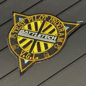 Könnte beinhalten: Gestickter Aufnäher mit dem Text "TEST PILOT PROGRAM" und "BATTLETECH" in einem kreisförmigen Design. Der Aufnäher ist schwarz mit goldenen und hellblauen Akzenten, und die Buchstaben "V.G.L." befinden sich unten.
