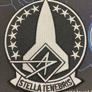 Puede incluir: Un parche bordado en blanco y negro con un diseño en forma de estrella y el texto "STELLA TENEBRIS".