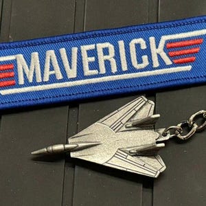 May include: A keychain featuring a blue fabric tag with the word "MAVERICK" in white, and red and white wing accents. A silver-colored fighter jet charm is attached to the keychain.