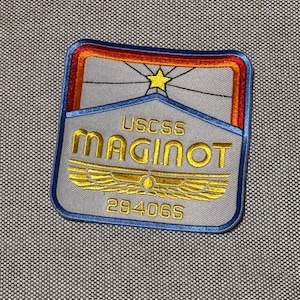 May include: Embroidered patch with the text "USCSS MAGINOT" in gold thread. The patch has a rectangular shape with a blue border and a star design at the top. The background is gray.