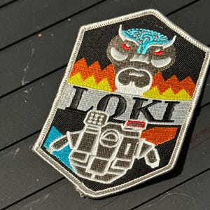 May include: A black, hexagonal patch with the word "LOKI" in silver lettering. The patch features a stylized owl-like face with blue and red accents, and a detailed illustration of a device below the text. The patch is bordered with white stitching.