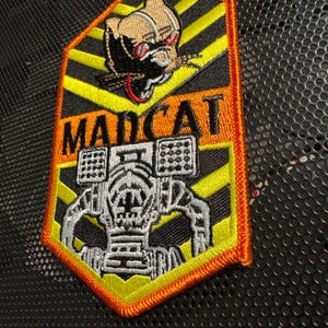 Może przedstawiać: Haftowana naszywka z napisem "MADCAT" i wzorem robota. Naszywka przedstawia głowę kota z czerwonymi oczami i robota o szczegółowym wzorze. Naszywka ma czarno-żółto-pomarańczową kolorystykę.