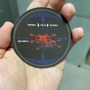 Puede incluir: Una pegatina circular en blanco y negro con un gráfico rojo y blanco de una nave espacial con el texto "FIRST STRIKE" y "THIS IS THE WAY" en la parte superior y "DEF-000-X" en la parte inferior.