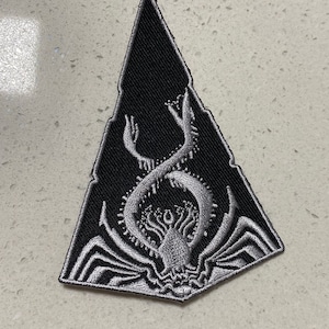 Op de afbeelding: Een zwarte driehoekige patch met een zilveren geborduurde afbeelding van een spinachtig wezen met meerdere poten en een gekrulde staart.