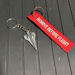 May include: A silver-colored metal airplane-shaped keychain with a chain and ring next to a red fabric tag with the words "REMOVE BEFORE FLIGHT" in white. The items are on a dark gray surface.