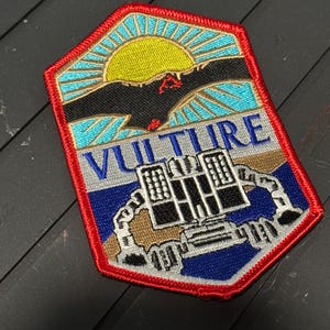 May include: Embroidered patch with a red border, featuring a stylized design. The patch includes a yellow sun, a black bird, and the word "VULTURE" in blue and silver. A building-like structure is at the bottom.