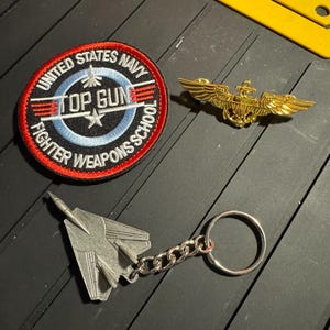 May include: A collection of Top Gun memorabilia, including a circular patch with the text "United States Navy Top Gun Fighter Weapons School", a gold-colored pilot's wings pin, and a silver-colored jet fighter keychain.
