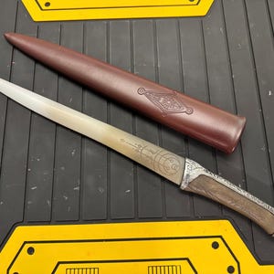 Könnte beinhalten: Ein silbernes Dolch mit einem Holzgriff und einer braunen Lederscheide. Die Klinge ist mit komplizierten Gravuren verziert und der Griff hat einen silbernen Ring.