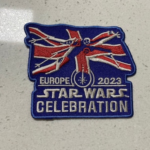 Puede incluir: Un parche azul bordado con un diseño de bandera Union Jack rojo, blanco y azul. La bandera está rodeada por un contorno blanco y el texto "EUROPE 2023 STAR WARS CELEBRATION" está impreso debajo de la bandera.