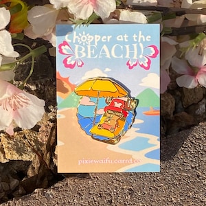 May include: A colorful enamel pin featuring a cartoon character relaxing on a beach chair under a yellow umbrella. The pin is set against a light blue background with the words "Chopper at the BEACH!" and pink butterfly accents. The pin is on a card with the website address pixiewaifu.carrd.co.