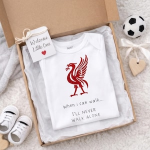 Può includere: Un body bianco per neonati con un emblema Liverbird rosso e la scritta "When I can walk... I'll never walk alone" all'interno di una confezione regalo. Un'etichetta recita "Welcome Little One". Una piccola palla da calcio e scarpine da bambino sono anche nell'immagine.