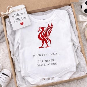 Puede incluir: Un body blanco para bebé con un emblema de pájaro rojo y el texto "When I can walk... I'll NEVER WALK ALONE" se presenta en una caja de regalo con un balón de fútbol, zapatos de bebé y un corazón de madera. Una etiqueta dice "Welcome Little One".