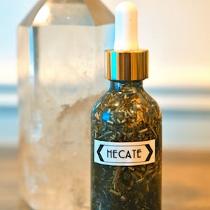 May include: A glass bottle with a gold lid and a dropper top. The bottle is filled with a dark liquid and herbs. The label on the bottle reads "HECATE".