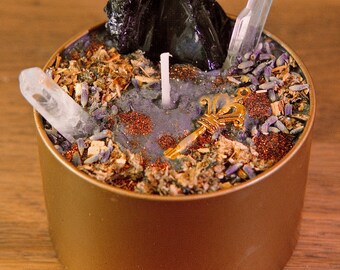 May include: A gold-colored candle tin filled with a purple wax base, dried herbs, and crystals. A black stone and two clear quartz crystals stand out. A gold-colored key charm and a white candle are also visible.