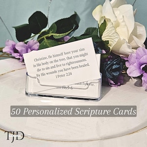 May include: A clear acrylic card holder filled with personalized scripture cards. The cards feature a Bible verse and the name "Christine." The holder sits on a white surface, surrounded by purple and white flowers. The text "50 Personalized Scripture Cards" is also visible.