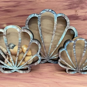 May include: Three decorative seashell-shaped boxes made of wood with a blue and brown paint finish. The boxes are open and have a clear glass insert. The smallest box is filled with small seashells.