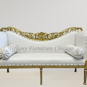 May include: White tufted chaise lounge with gold ornate scrollwork and legs. The chaise has two white cylindrical pillows with button tufting. The text "Tiffany Furniture Chicago" is visible on the chaise.