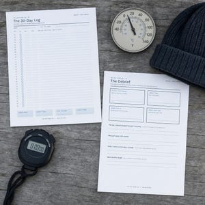Op de afbeelding: Twee witte papieren logbladen getiteld The 30-Day Log en The Debrief met zwarte tekst en lijnen, een ronde zilveren analoge thermometer met Celsius- en Fahrenheit-schalen, een zwarte digitale stopwatch en een donkergrijze gebreide muts.