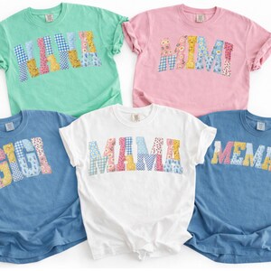 May include: Five short-sleeved t-shirts in various colors: mint green, pink, white, and two shades of blue. Each shirt features a different name in a patchwork design: MAMA, MIMI, GIGI, MAMA, and MEMAW. The names are in a colorful, patterned font.