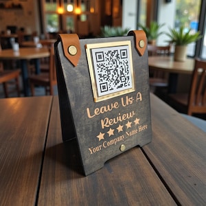 May include: A dark wooden table tent with a QR code and the text "Leave Us A Review". The sign has a gold border around the QR code and leather accents with brass rivets. The text "Your Company Name Here" is also displayed.