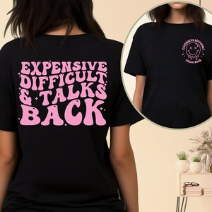 May include: Black t-shirt with pink text on the back that reads "Expensive Difficult & Talks Back". The front of the shirt has a pink graphic of a melting smiley face with the text "Expensive Difficult Talks Back".