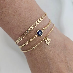 May include: Four gold bracelets stacked on a wrist. The top bracelet is a thick, gold chain. The second bracelet is a thinner gold chain with a blue evil eye charm. The third bracelet is a delicate gold chain with a small gold letter 'M' charm. The bottom bracelet is a thin gold chain with small gold beads.