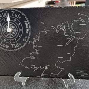 Puede incluir: Un reloj de pizarra negra con un gráfico de mareas y una brújula. La esfera del reloj muestra las palabras "High Tide" y "Low Tide" con una aguja blanca que apunta al nivel de marea actual. El gráfico muestra la costa de Irlanda con varias ciudades y pueblos etiquetados.