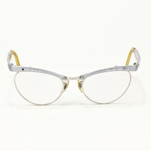 May include: Silver-framed cat-eye glasses with clear lenses. The frames have a decorative design along the top edge and yellow earpieces. The glasses are set against a white background.