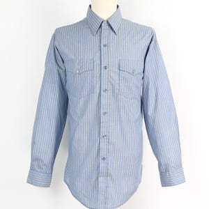 Può includere: Camicia azzurra a maniche lunghe con righe verticali gialle e bianche. La camicia ha due tasche sul petto, una chiusura con bottoni e polsini abbottonati. La camicia è esposta su un manichino.