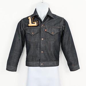 May include: Dark wash denim jacket with a patch featuring a yellow and navy blue letter "L". The jacket has two chest pockets, button closures, and orange stitching. The jacket is displayed on a white mannequin.