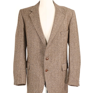 Puede incluir: Un blazer de tweed marrón con un patrón de espiga. La chaqueta tiene solapas con muescas, dos bolsillos delanteros y dos botones marrones. El blazer se muestra en un maniquí.