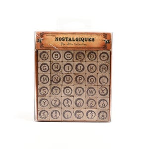 May include: A collection of wooden alphabet stamps in a clear plastic case. The stamps are arranged in a grid and feature letters A to Z, along with numbers and symbols. The packaging reads "NOSTALGIQUES The Attie Collection".