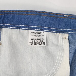 May include: Close-up of blue denim jeans with a white pocket and a tag. The tag reads "MADE IN HONDURAS OF U.S. FABRIC" and lists the fabric composition, size 106.68 x 73.66 cm, and a product code.
