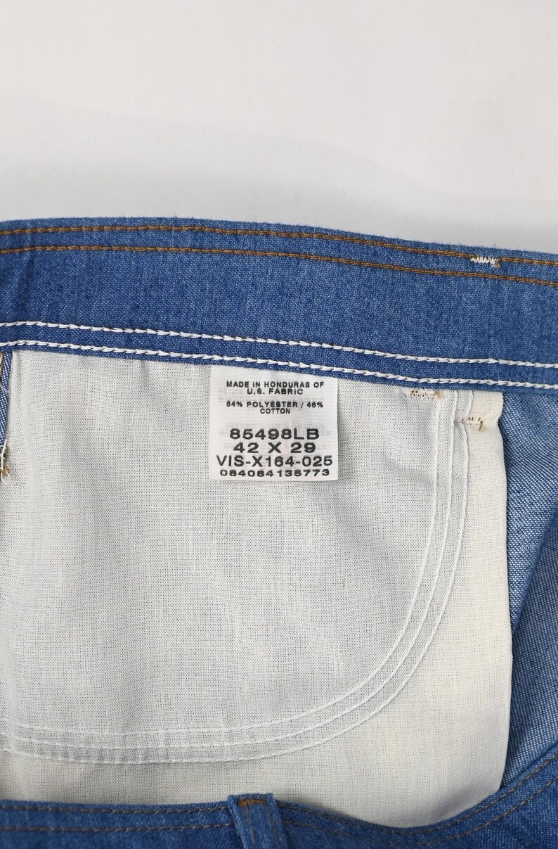 May include: Close-up of blue denim jeans with a white pocket and a tag. The tag reads "MADE IN HONDURAS OF U.S. FABRIC" and lists the fabric composition, size 106.68 x 73.66 cm, and a product code.