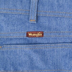 K&ouml;nnte beinhalten: Nahaufnahme einer hellblauen Jeans mit sichtbaren N&auml;hten und einem braunen Wrangler-Etikett auf der Ges&auml;&szlig;tasche. Die Jeans hat ein klassisches Design mit G&uuml;rtelschlaufe und Tasche.