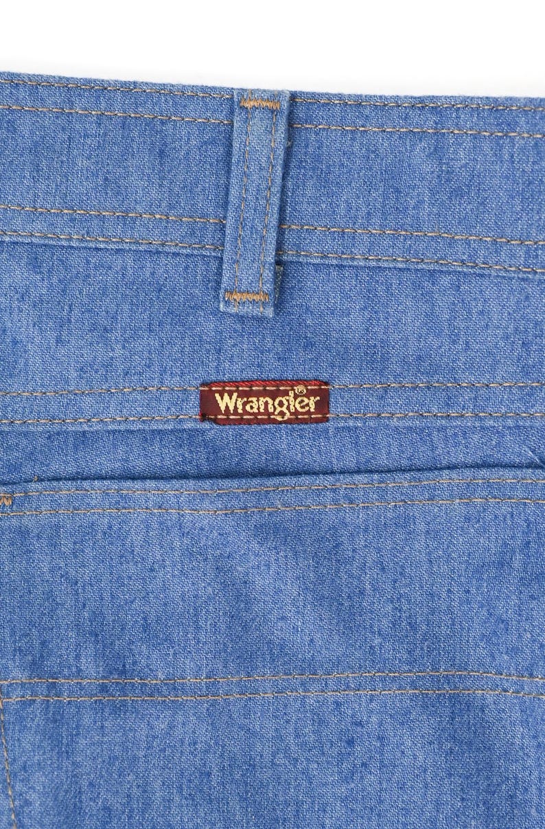 K&ouml;nnte beinhalten: Nahaufnahme einer hellblauen Jeans mit sichtbaren N&auml;hten und einem braunen Wrangler-Etikett auf der Ges&auml;&szlig;tasche. Die Jeans hat ein klassisches Design mit G&uuml;rtelschlaufe und Tasche.