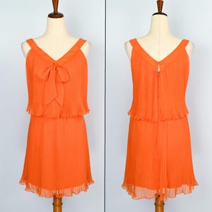 May include: An orange pleated dress with a V-neckline and a bow detail. The dress has a sleeveless design and a flowing skirt. The dress is displayed on a mannequin. The dress is a vibrant orange color.