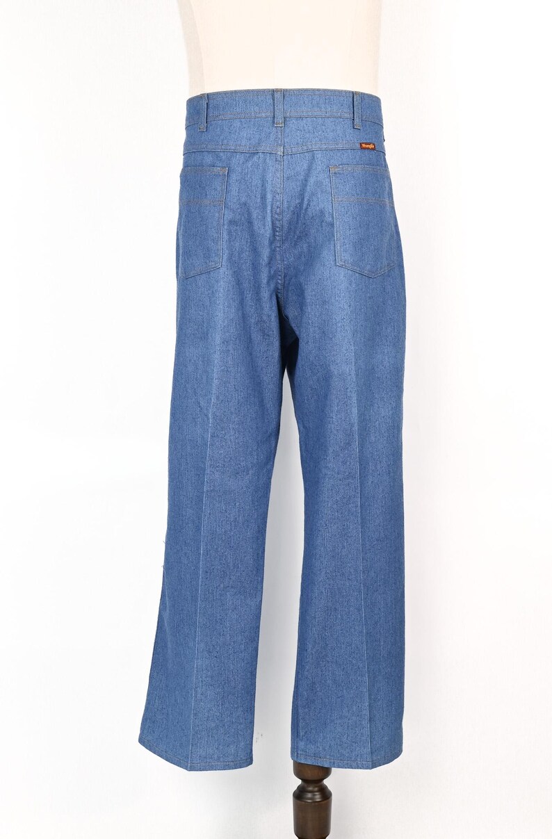 May include: Light blue denim jeans with a classic five-pocket design. The jeans have a straight leg cut and a high waist. The back pockets are square. The Wrangler logo is visible on the back pocket.