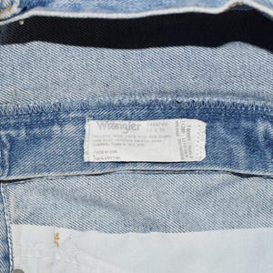 Peut inclure: Gros plan sur une ceinture de jean bleu clair avec une &eacute;tiquette Wrangler blanche. L'&eacute;tiquette porte le texte "Wrangler" en caract&egrave;res gras et comprend des instructions d'entretien et des informations sur la taille. L'&eacute;tiquette est fix&eacute;e &agrave; l'int&eacute;rieur de la ceinture.