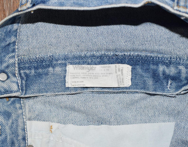 Peut inclure: Gros plan sur une ceinture de jean bleu clair avec une &eacute;tiquette Wrangler blanche. L'&eacute;tiquette porte le texte "Wrangler" en caract&egrave;res gras et comprend des instructions d'entretien et des informations sur la taille. L'&eacute;tiquette est fix&eacute;e &agrave; l'int&eacute;rieur de la ceinture.