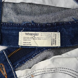 以下が含まれることがあります： Wranglerのジーンズの内側のクローズアップ。ラベルには「Wrangler 31 MWZW 30 x 34」と「Made in USA 100% Cotton」と記載されています。ジーンズは、白いステッチとジッパーが付いた濃い青色のデニムです。
